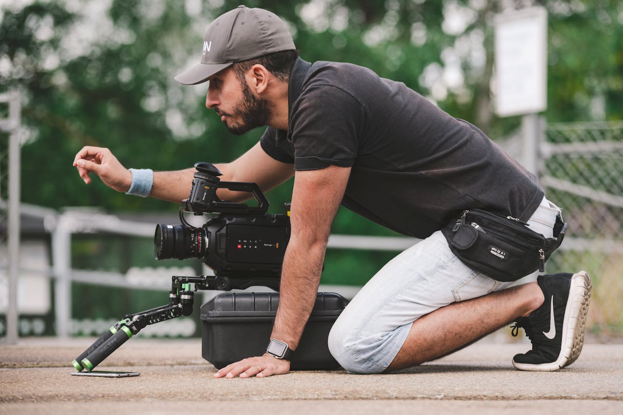 A professional cinematographer kneeling with camera equipment outdoors, capturing a scene in daylight.